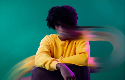 Woman with dark curls in a yellow sweater sitting in front of a plain green background. Her face has merged into a long blur of colour.