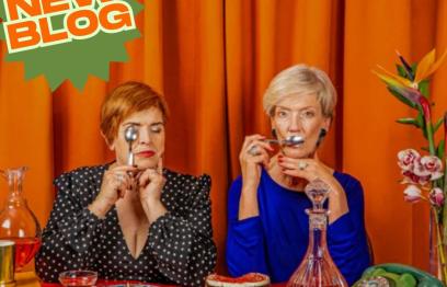 2 women sitting at a table with cake and wine in front of an orange curtain