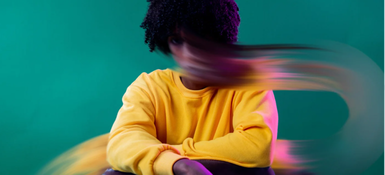 Woman with dark curls in a yellow sweater sitting in front of a plain green background. Her face has merged into a long blur of colour.