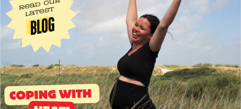 A pregnant woman smiling and waving her arms in the air. She is standing among sand dunes.