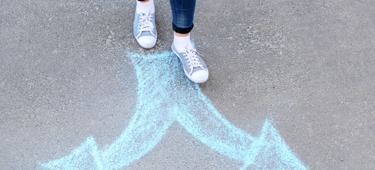 Image of a person's legs and feet in jeans and sneakers taking steps towards two chalk drawn arrows.